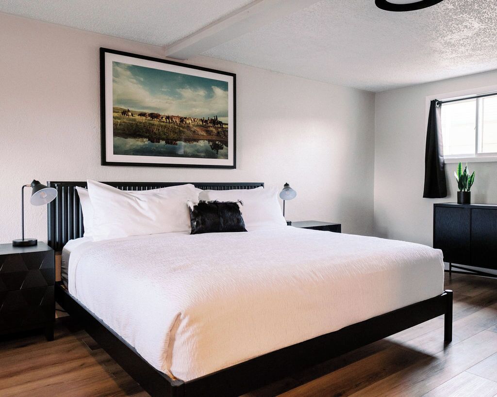 A modern bedroom with a large bed, a framed landscape painting, a nightstand with a lamp, and a window with a view.