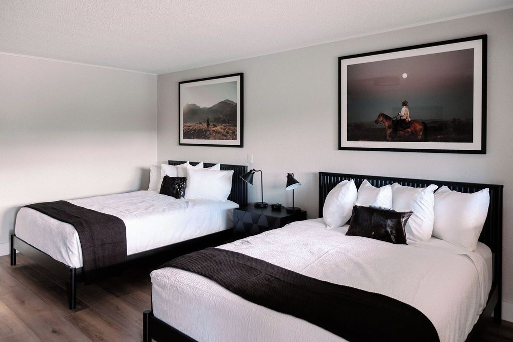 Een hotelkamer met twee bedden, elk voorzien van witte lakens en zwarte hoofdborden. Twee ingelijste foto's hangen aan de muur, waarvan er een een woestijnlandschap toont en de andere een ruiter.