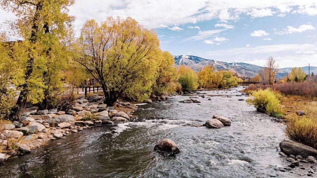 Un río con rocas y árboles, un puente y montañas al fondo.