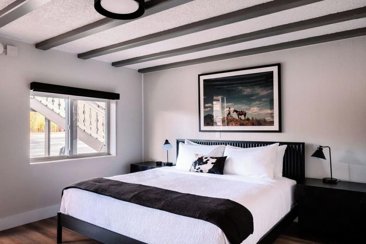 A modern bedroom with a large bed, a window with a view of buildings, a framed picture on the wall, and a ceiling with exposed beams.
