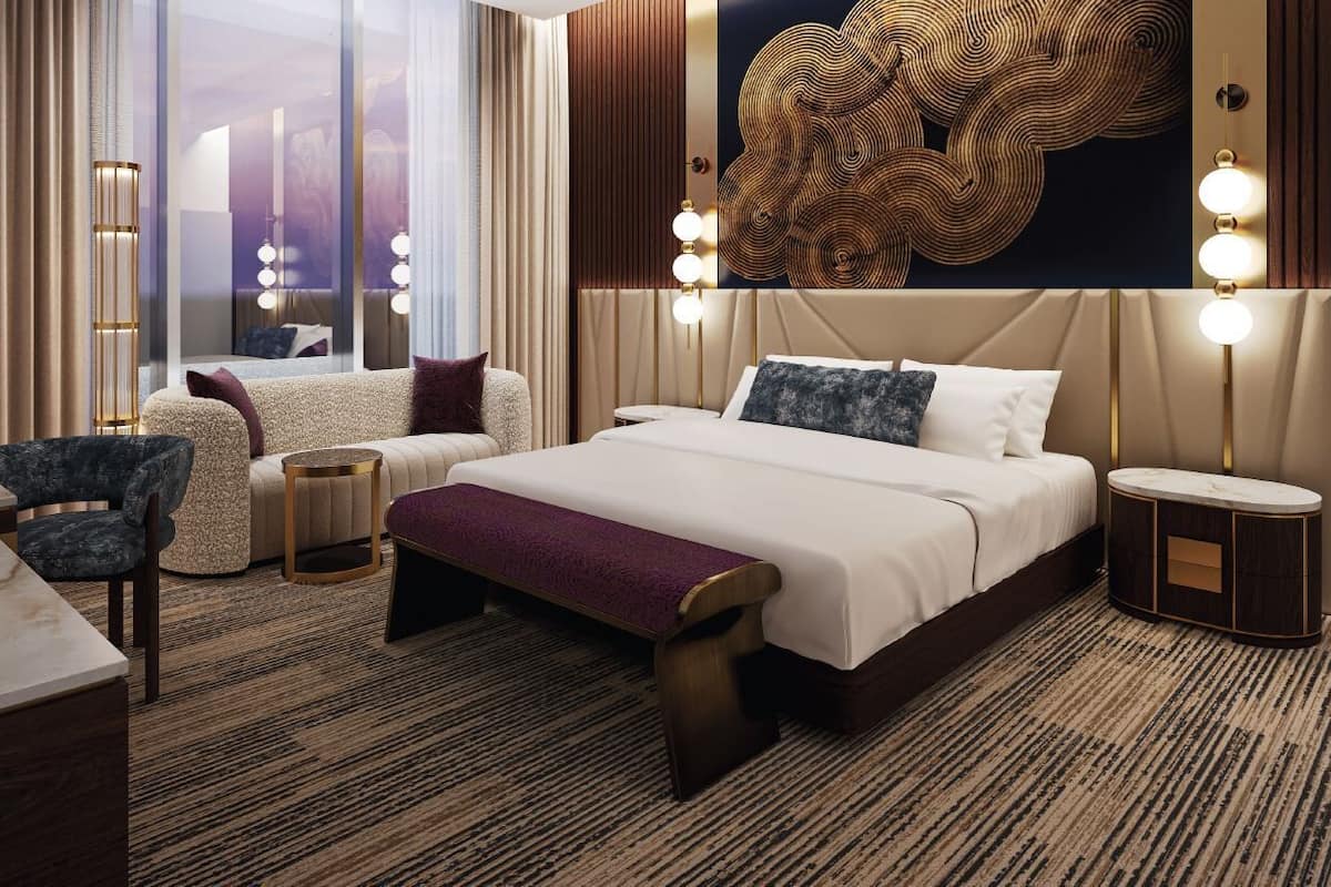 Une chambre d’hôtel moderne dotée d’un grand lit, d’un coin salon avec un fauteuil et une petite table, ainsi que d’une grande fenêtre avec des rideaux.