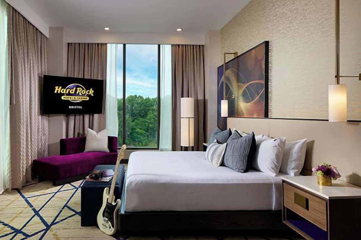 Une chambre d’hôtel avec un lit, une chaise, une guitare, un téléviseur diffusant des images de l’hôtel et du casino « Hard Rock », et une fenêtre donnant sur des arbres.
