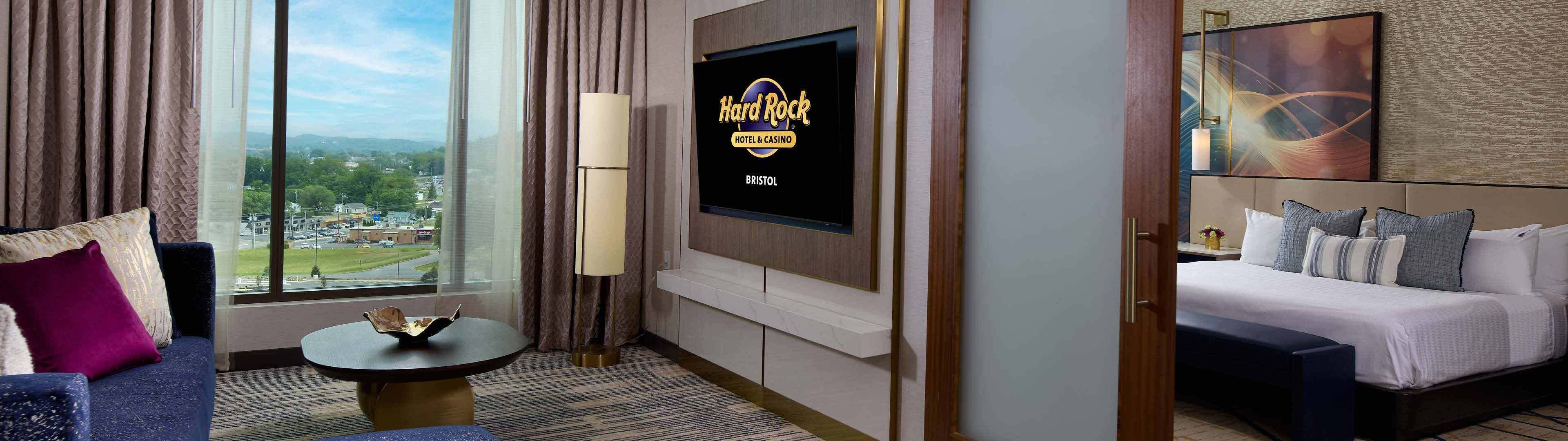 A hotel room with a large window offering a view of a cityscape, a flat-screen TV mounted on the wall, a bed with multiple pillows, and a small round table with a lamp.