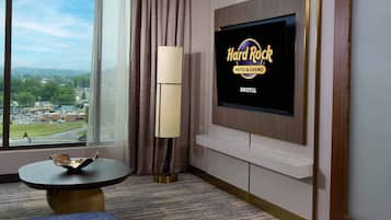 A hotel room with a large window offering a view of a cityscape, a flat-screen TV mounted on the wall, a bed with multiple pillows, and a small round table with a lamp.