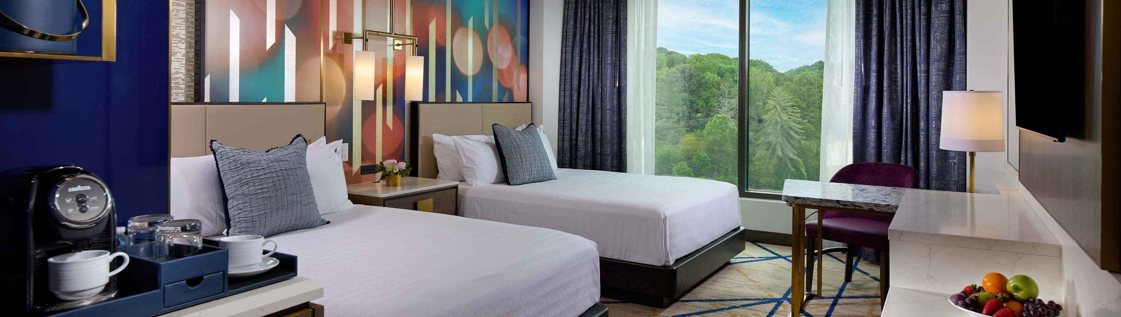 Quarto de hotel com duas camas, uma escrivaninha com abajur, máquina de café e vista para uma floresta.