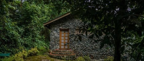 A stone building with a wooden door, surrounded by lush greenery and a stone pathway.