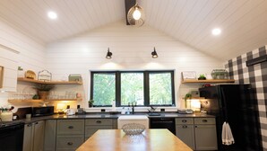 Une cuisine avec une table en bois, deux lampes suspendues, un réfrigérateur noir et une fenêtre donnant sur des arbres.