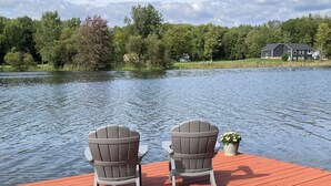 Une terrasse en bois avec deux fauteuils Adirondack donnant sur un lac, entourée d’arbres et avec une maison en arrière-plan.