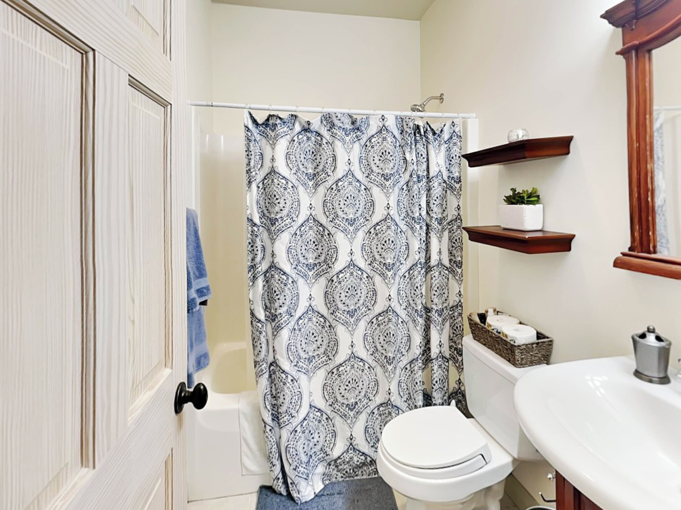 A bathroom with a patterned shower curtain, a white toilet, a sink with a mirror, and wooden shelves with items.