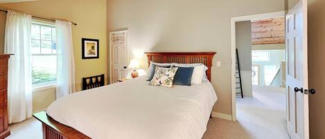 A bedroom with a large bed, a wooden headboard, a bedside table with a lamp, a chair, and a door leading to a staircase.