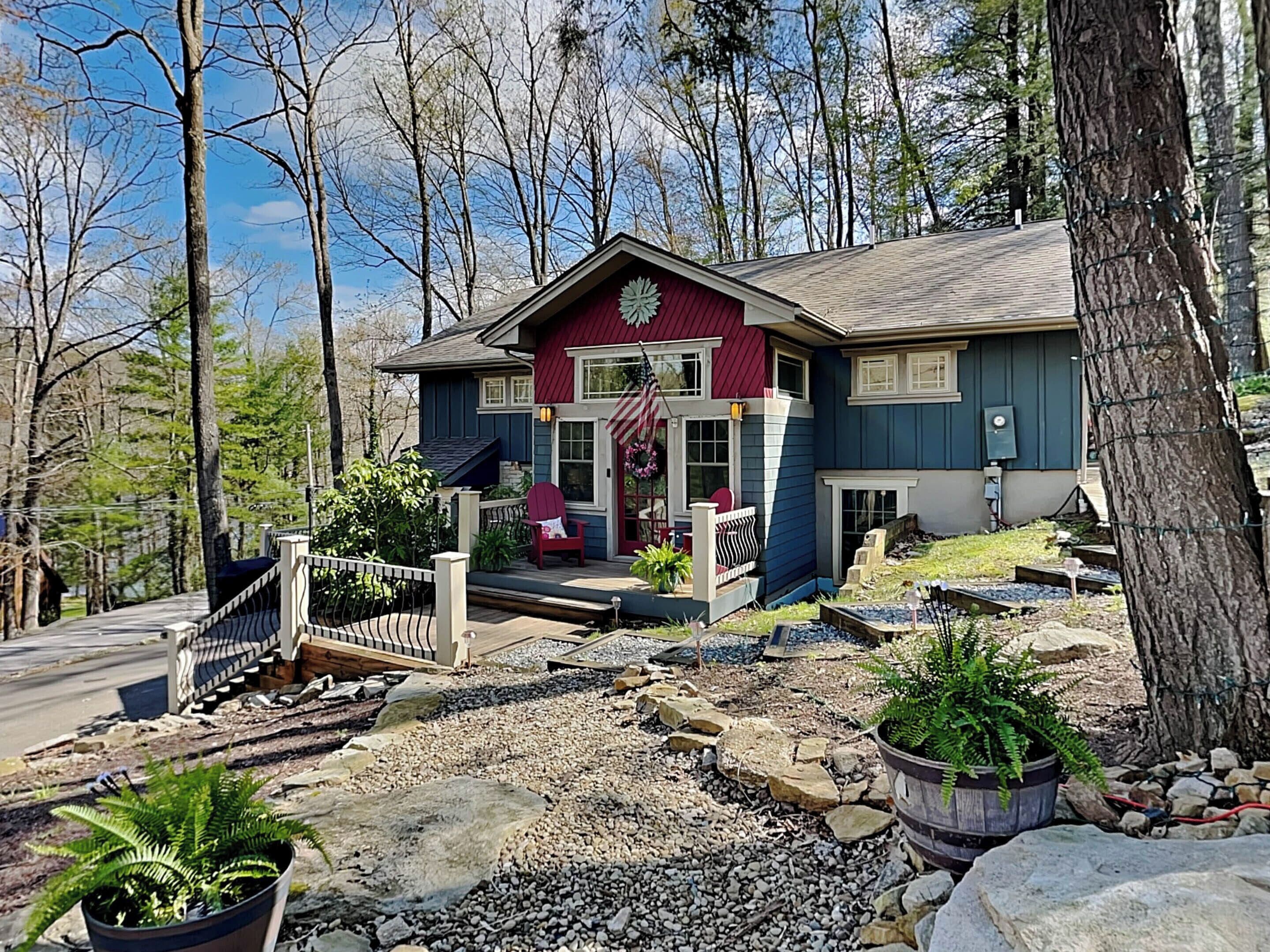 A two-story house with a red front door, a porch with a red chair, and a small garden with rocks and plants.