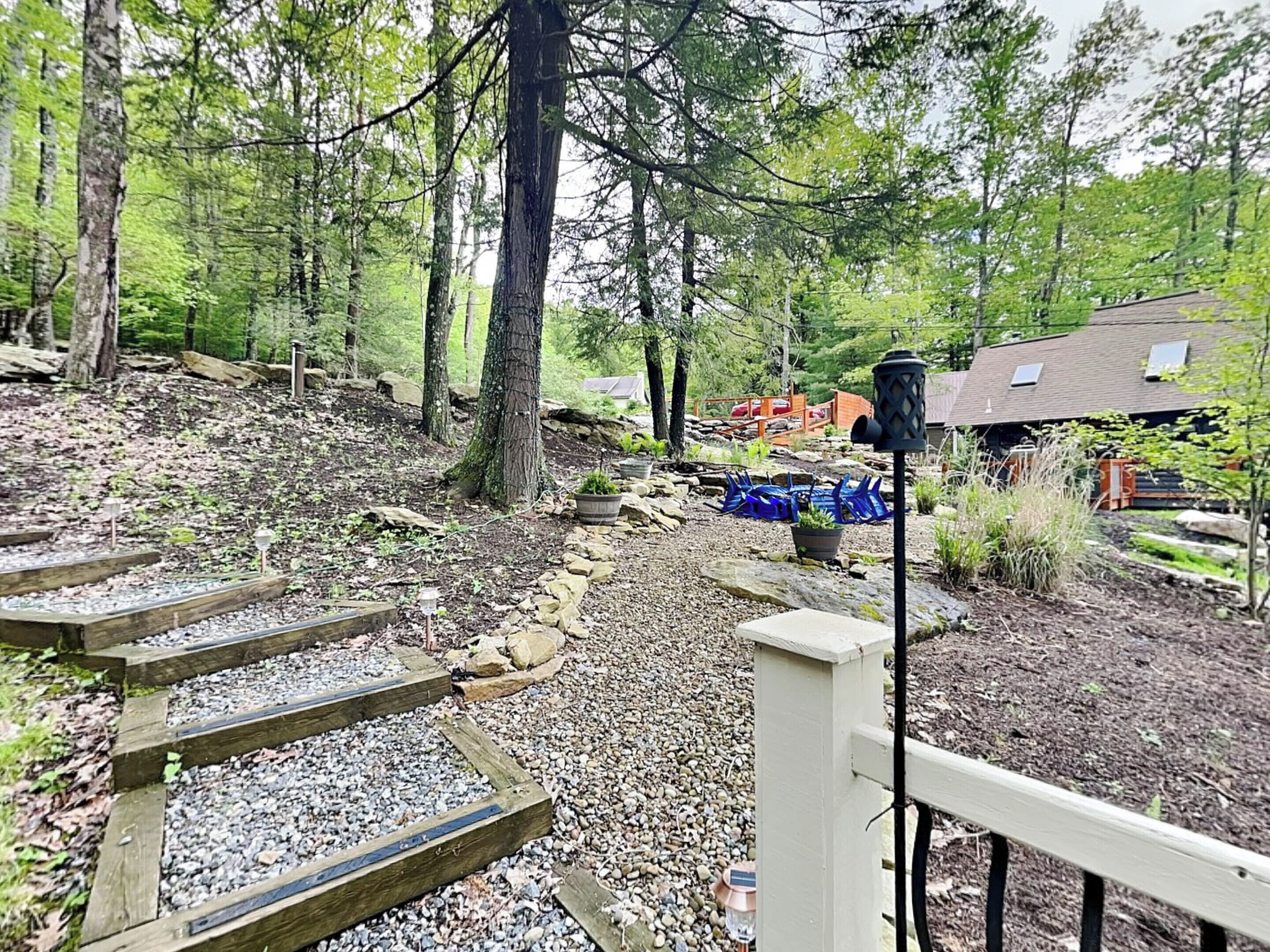 A wooden staircase leading up a gravel path surrounded by trees, with a house and outdoor seating area in the background.