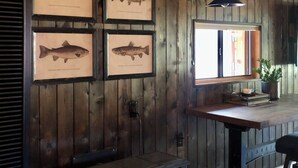 Un intérieur rustique avec des murs en bois, des illustrations de poissons encadrées, un poêle d’époque et une fenêtre offrant une belle vue.