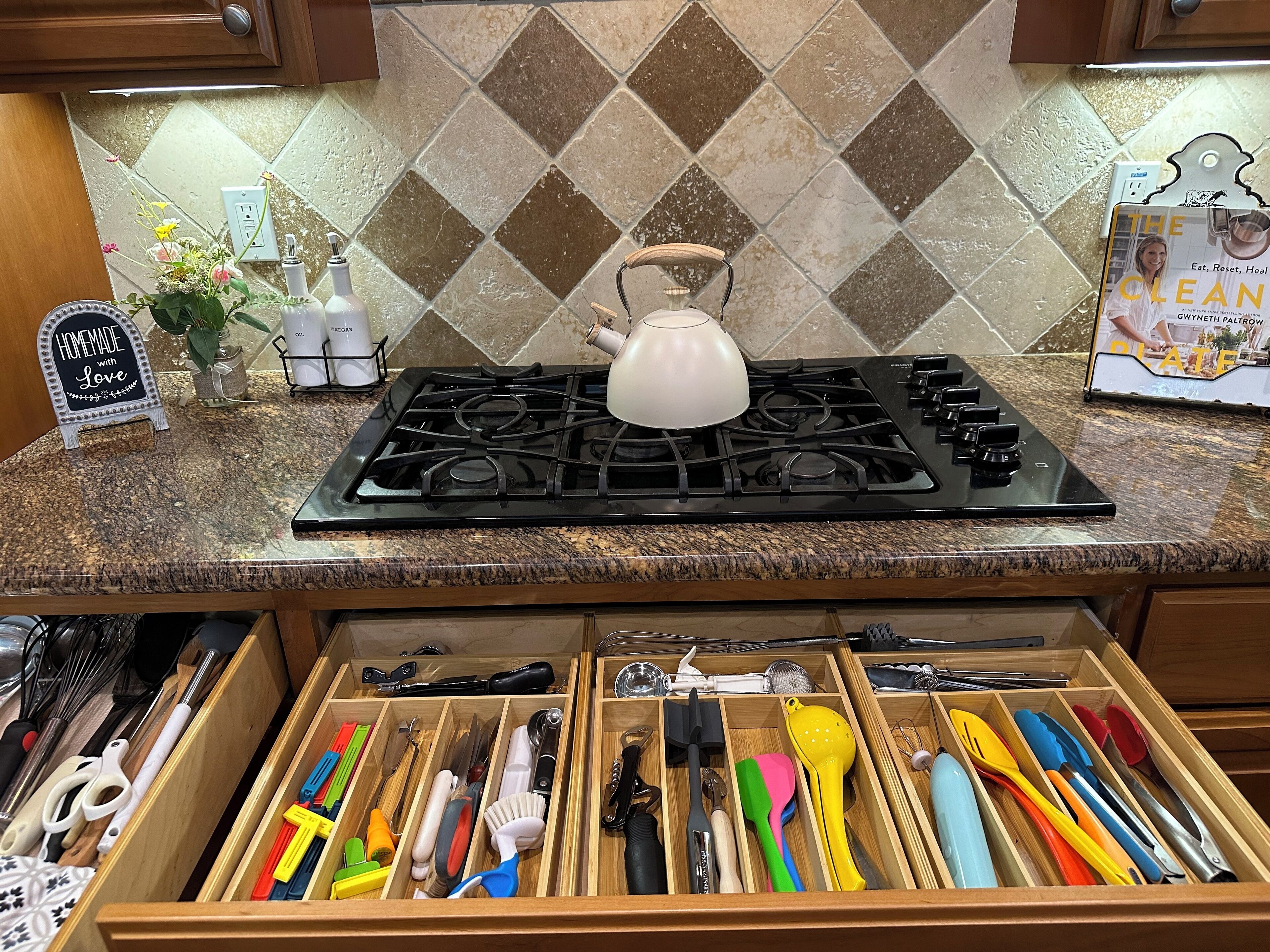 A kitchen with a gas stove, a kettle, and a variety of utensils in a drawer.
