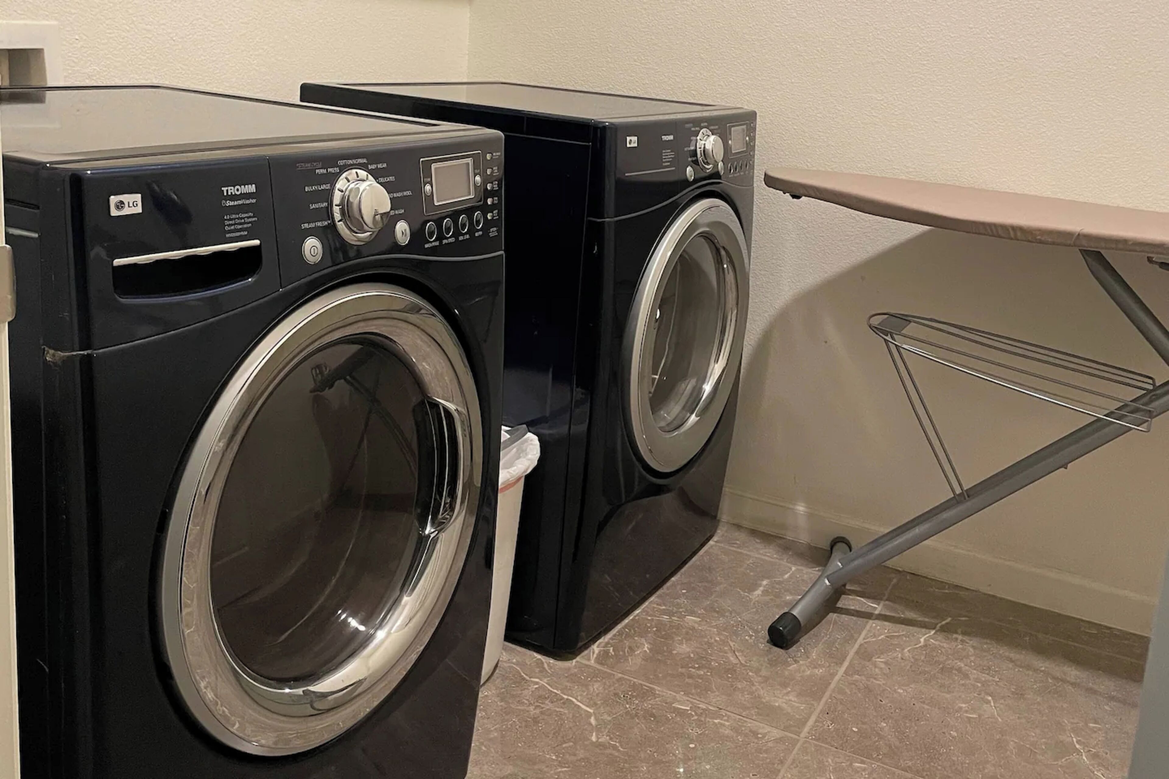 Un lave-linge noir de la marque LG, modèle TROMM, doté d’un panneau de commande et d’une porte transparente, placé à côté d’un étendoir mural.