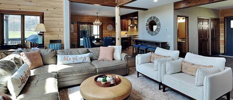 A spacious living room with a sectional sofa, a round coffee table, and a white sofa. A kitchen is visible in the background.