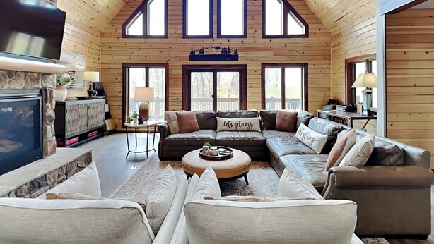Un intérieur de chalet chaleureux avec une cheminée en pierre, des murs en bois, un grand canapé d’angle et un plafond haut doté de fenêtres de toit.