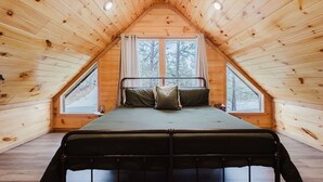 Un dormitorio acogedor en el ático con techo inclinado, una cama con estructura metálica y dos ventanas con vistas a árboles.