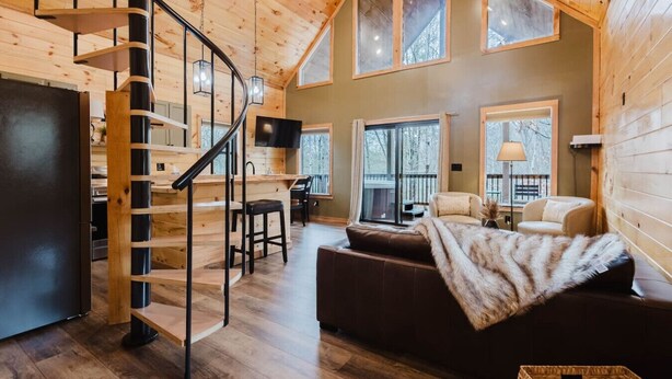 Un interior de cabaña espacioso con escalera de madera, una zona de estar acogedora y grandes ventanales con vistas al exterior.