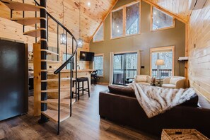 A spacious cabin interior with a wooden staircase, a cozy seating area, and large windows offering views of the outdoors. - Timber Pines at Red River Gorge (Zoe)