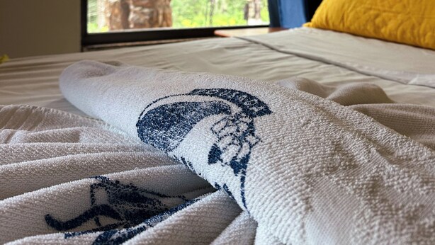 A bed with a white and blue towel, a yellow pillow, and a view of a tree through a window. - Pousada Sanhaço (Angra dos Reis)