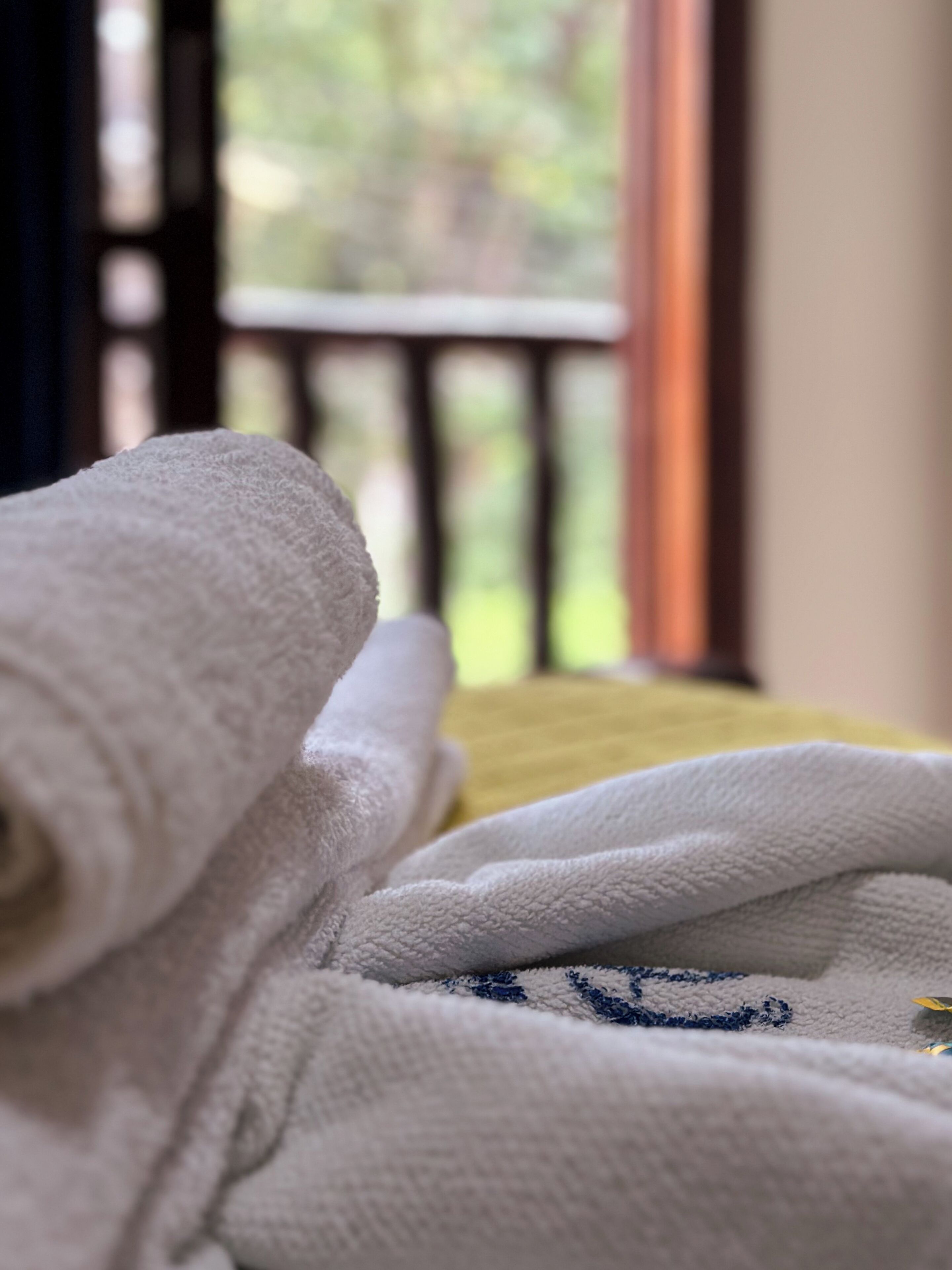 Primer plano de una toalla blanca con un diseño azul bordado, sobre una superficie amarilla, posiblemente una cama o sofá, con una vista borrosa de un balcón y follaje al exterior.