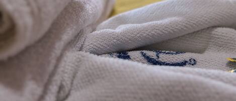 Primer plano de una toalla blanca con un diseño azul bordado, sobre una superficie amarilla, posiblemente una cama o sofá, con una vista borrosa de un balcón y follaje al exterior.