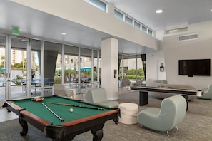 A pool table, pool cues, and chairs in a modern indoor space with large windows and a flat-screen TV. - Oceanside | Coastal Vibes | Pool & Hot Tub (Oceanside)