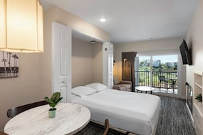A hotel room with a bed, a round table with a plant, a balcony with a table and chairs, and a TV mounted on the wall. - Oceanside | Coastal Vibes | Pool & Hot Tub (Oceanside)