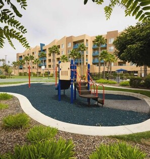 A playground with a climbing structure, surrounded by a landscaped area with palm trees and a building in the background.