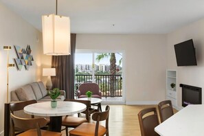 Un salon moderne avec une table à manger ronde, des chaises, un canapé, un téléviseur et un balcon offrant une vue sur les immeubles et les palmiers.