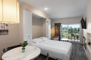 A hotel room with a bed, a round table with a plant, a balcony with a table and chairs, and a TV mounted on the wall. - Oceanside | Sunset Coast | Pool & Hot Tub (Oceanside)