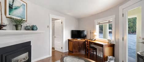 Una sala de estar con chimenea, televisor, escritorio con silla y vista a una masa de agua a través de una ventana.