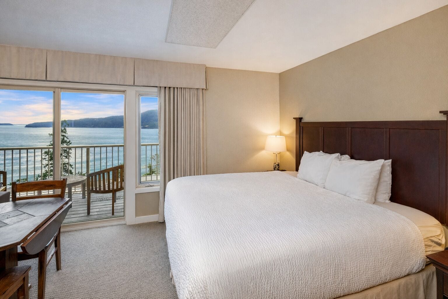 Habitación de hotel con una cama grande, un escritorio y una silla. Tiene un balcón con vista al agua y a las montañas.