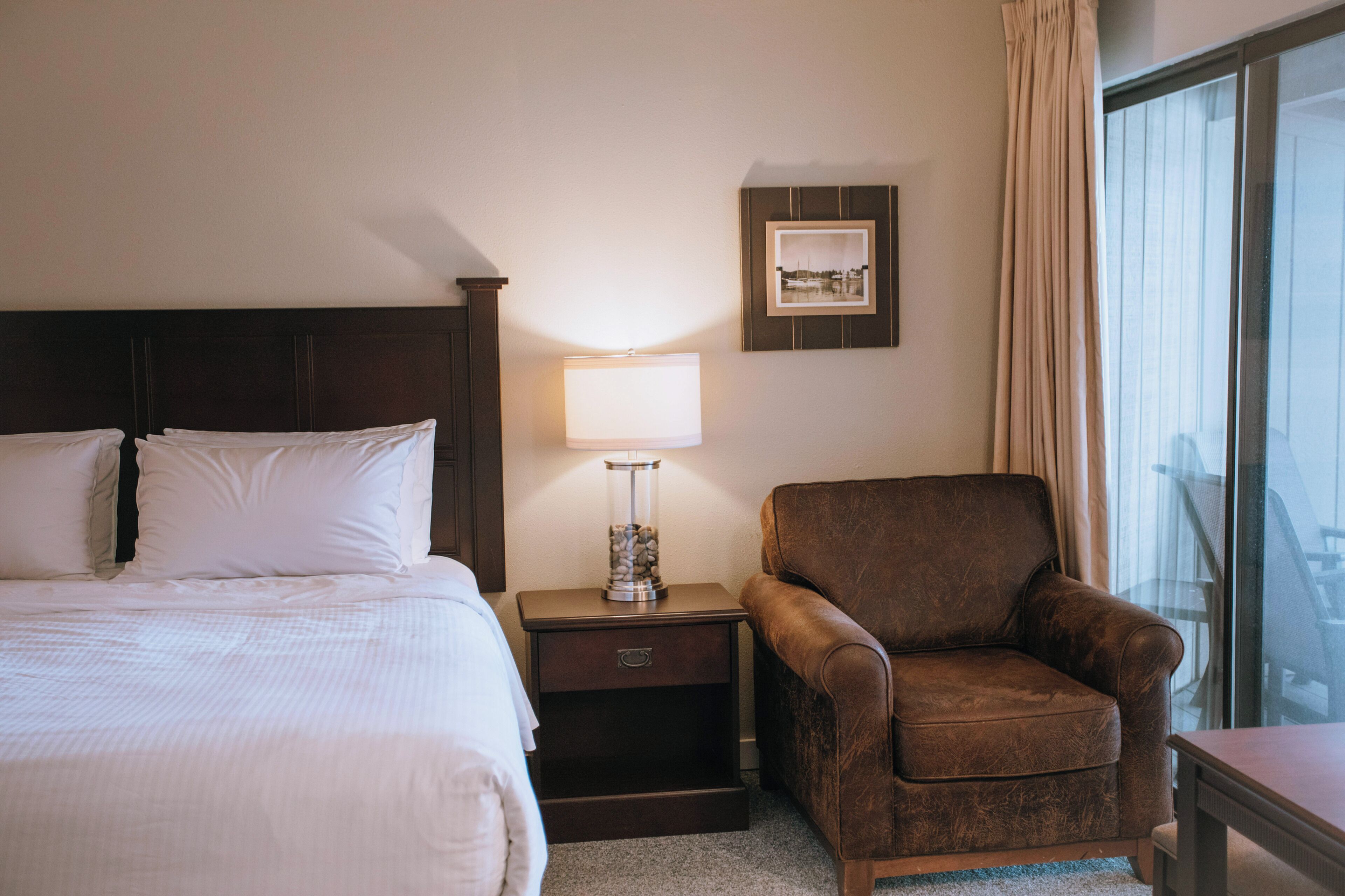 Een hotelkamer met een bed, een nachtkastje met een lamp, een stoel, een ingelijst schilderij aan de muur en een raam met gordijnen.
