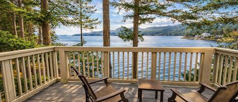 A wooden deck with two chairs and a table, overlooking a lake with trees and mountains in the background.