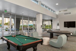 A pool table, pool cues, and chairs in a modern indoor space with large windows and a flat-screen TV.