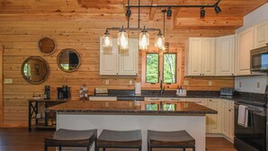 A kitchen with wooden walls, a central island with bar stools, a microwave, an oven, and a window with a view of trees. - Luna Stella Lodge-6BR Mt View, Hot Tub, Fire Pit (McHenry)