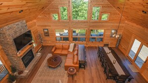 A spacious log cabin interior with a fireplace, leather seating, a dining area, and large windows offering views of the outdoors. - Luna Stella Lodge-6BR Mt View, Hot Tub, Fire Pit (McHenry)