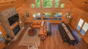 A spacious log cabin interior with a fireplace, leather seating, a dining area, and large windows offering views of the outdoors.