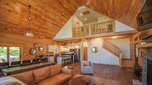 A spacious living area with a high ceiling, wooden beams, a central seating area with a sofa and armchairs, a fireplace, and a kitchen in the background. - Luna Stella Lodge-6BR Mt View, Hot Tub, Fire Pit (McHenry)