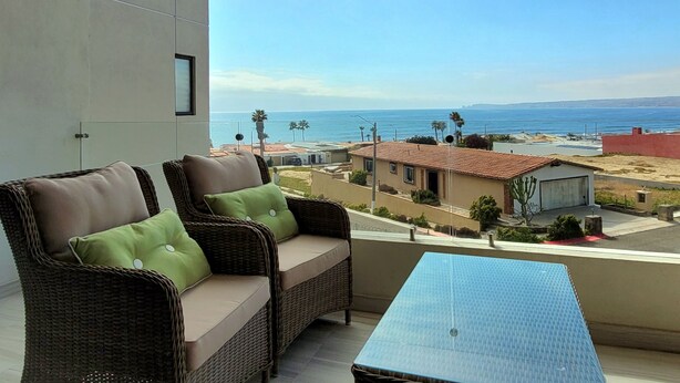 A balcony with wicker furniture and a glass table, overlooking a coastal area with houses and palm trees. - Lovely Condo with Stunning Ocean View (Playas de Rosarito)