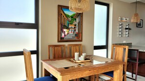 A dining area with a wooden table, blue cushioned chairs, a woven pendant light, and a painting above the table. - Lovely Condo with Stunning Ocean View (Playas de Rosarito)