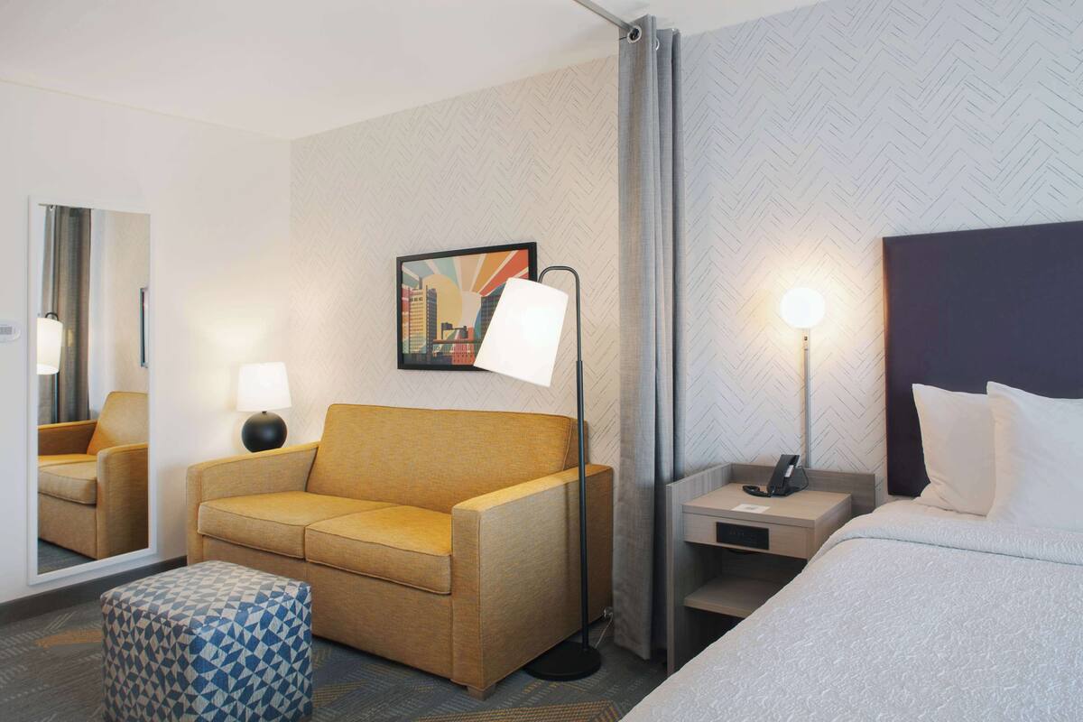 A hotel room with a sofa, a bed, a nightstand, a lamp, a mirror, and a cityscape painting on the wall.