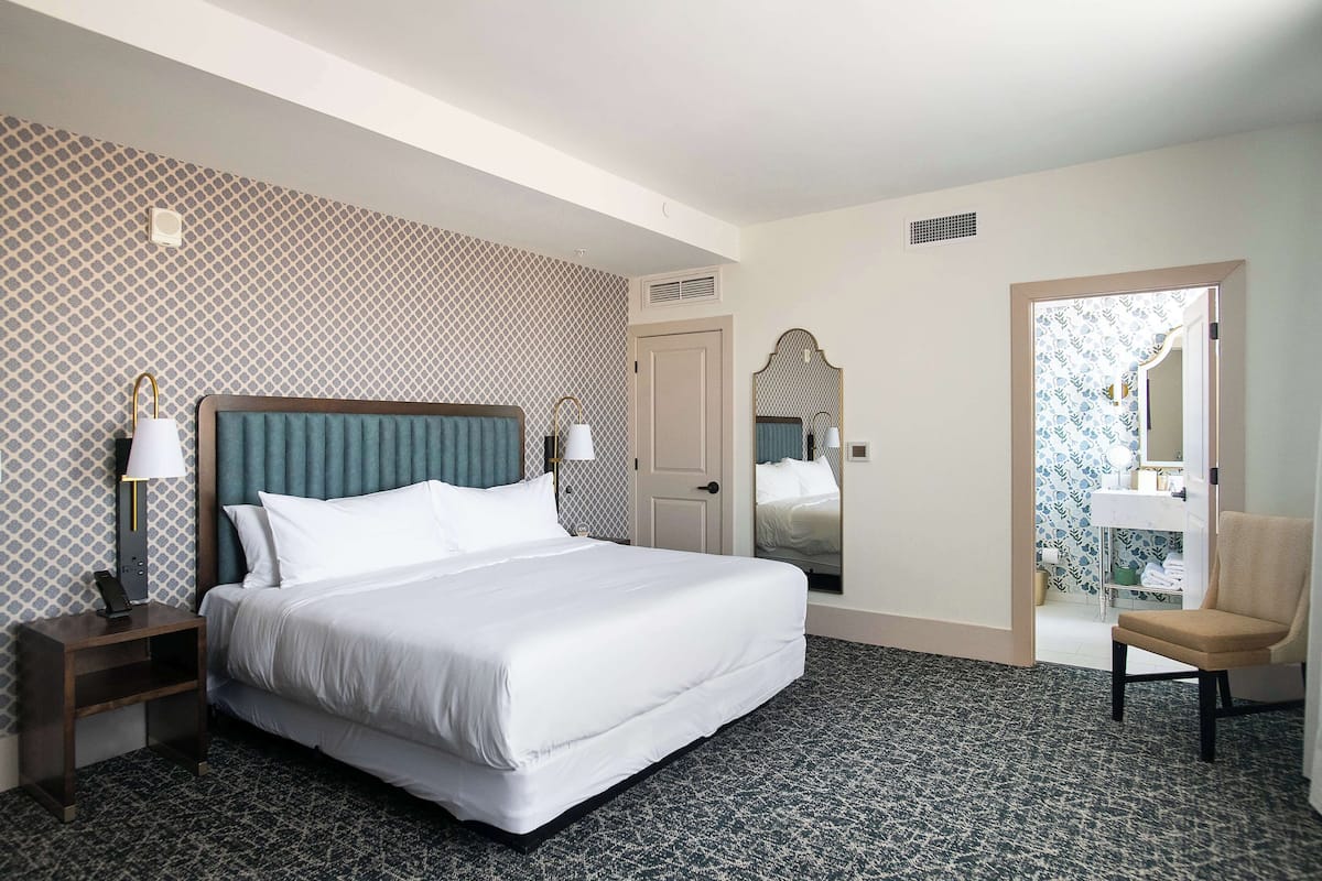 Quarto de hotel com uma cama grande, duas mesas de cabeceira com luminárias, uma cadeira e um banheiro visível por uma porta aberta.