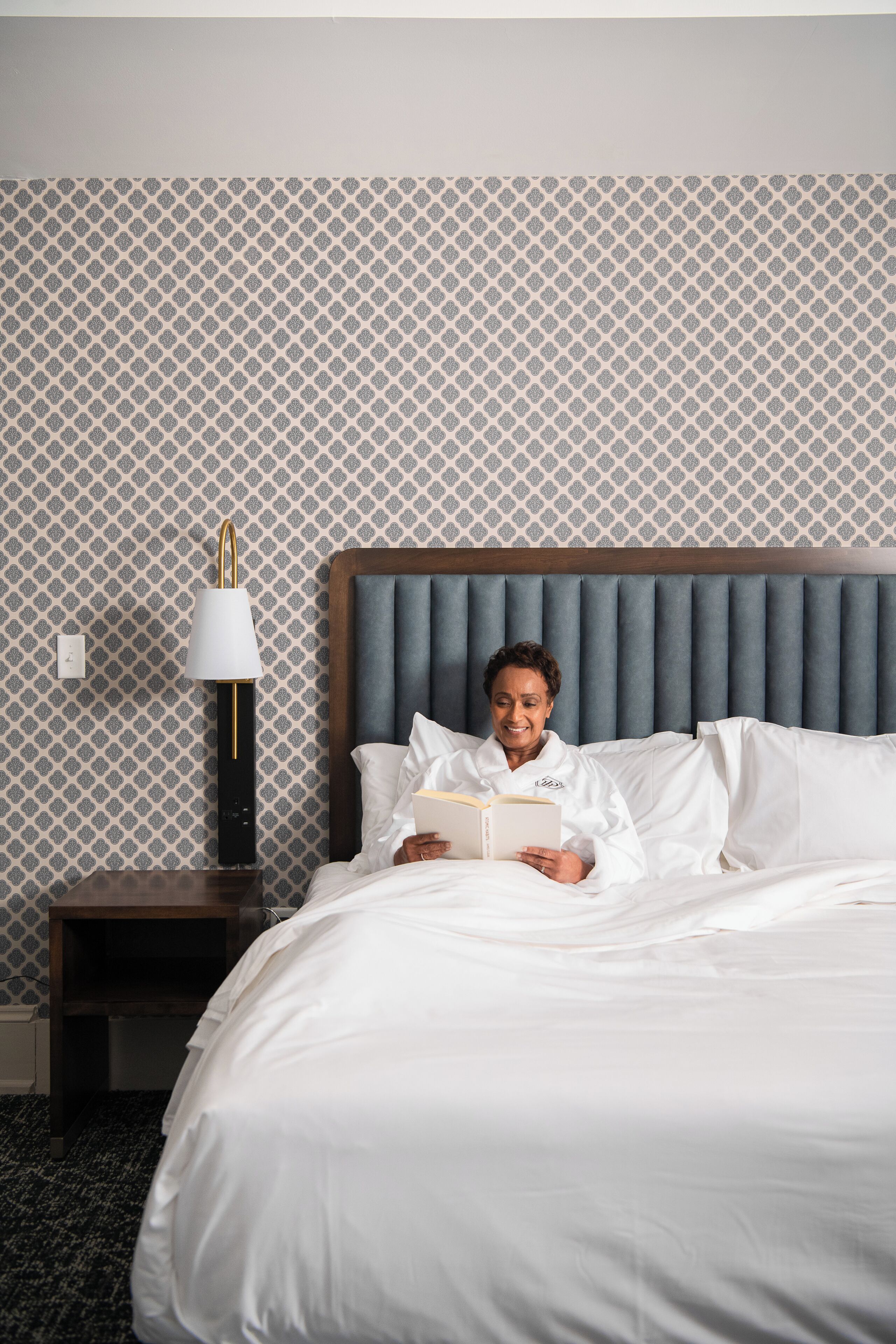 A person reading a book in a hotel room with a patterned wallpaper, a wooden headboard, and a nightstand with a lamp.