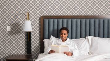 A person reading a book in a hotel room with a patterned wallpaper, a wooden headboard, and a nightstand with a lamp.