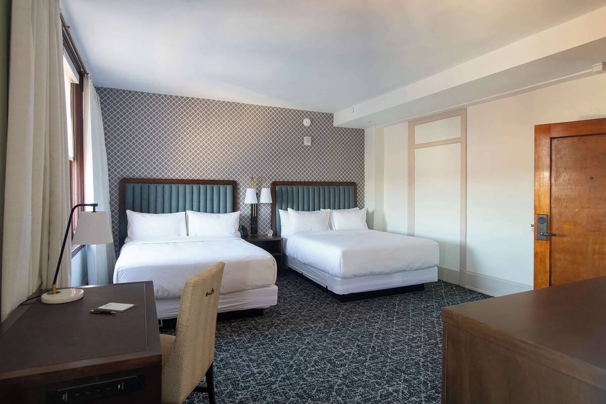Quarto de hotel com duas camas, uma escrivaninha, uma cadeira e uma porta de madeira.