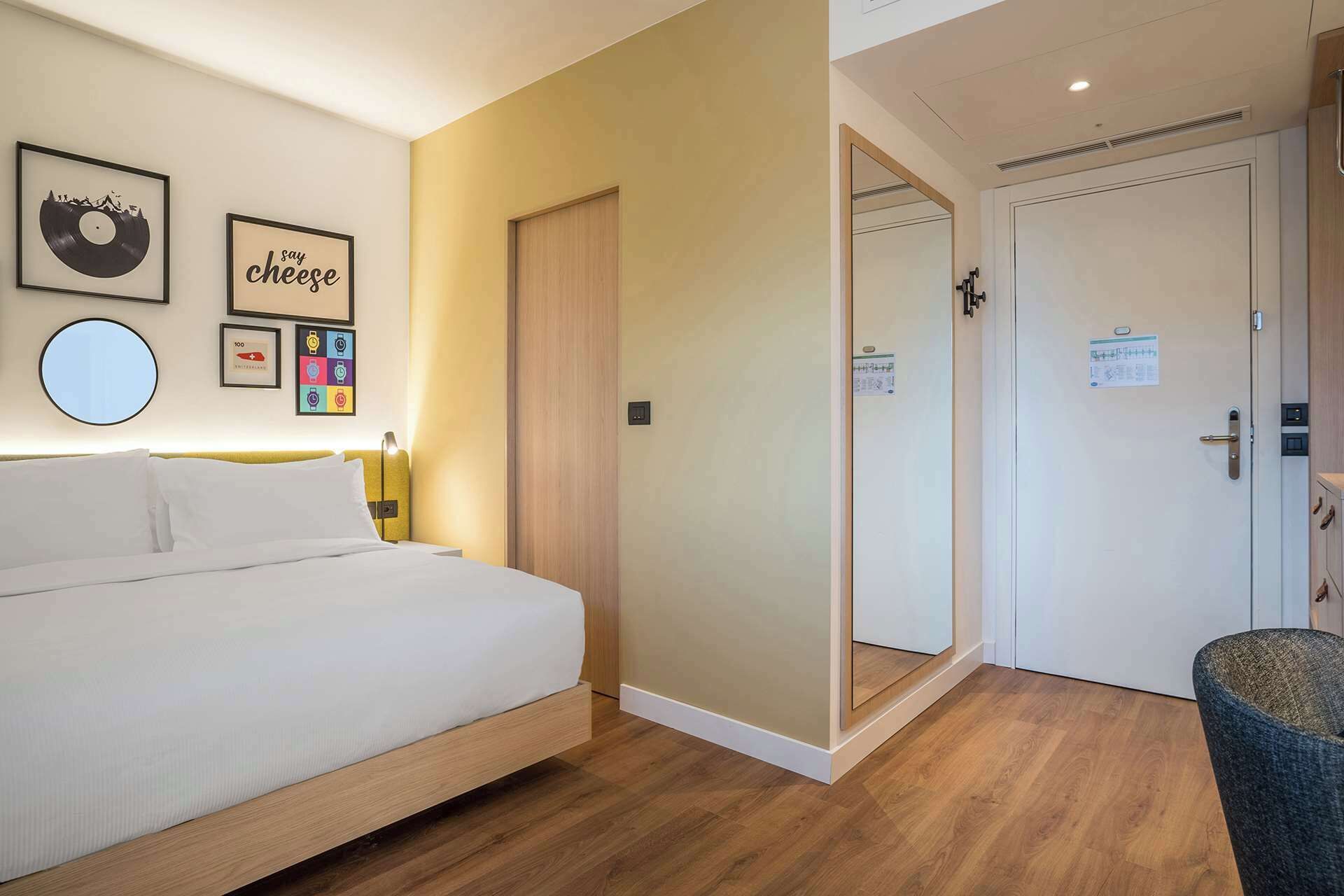 Quarto de hotel com cama, mesa de cabeceira, espelho, guarda-roupa e porta com uma placa.