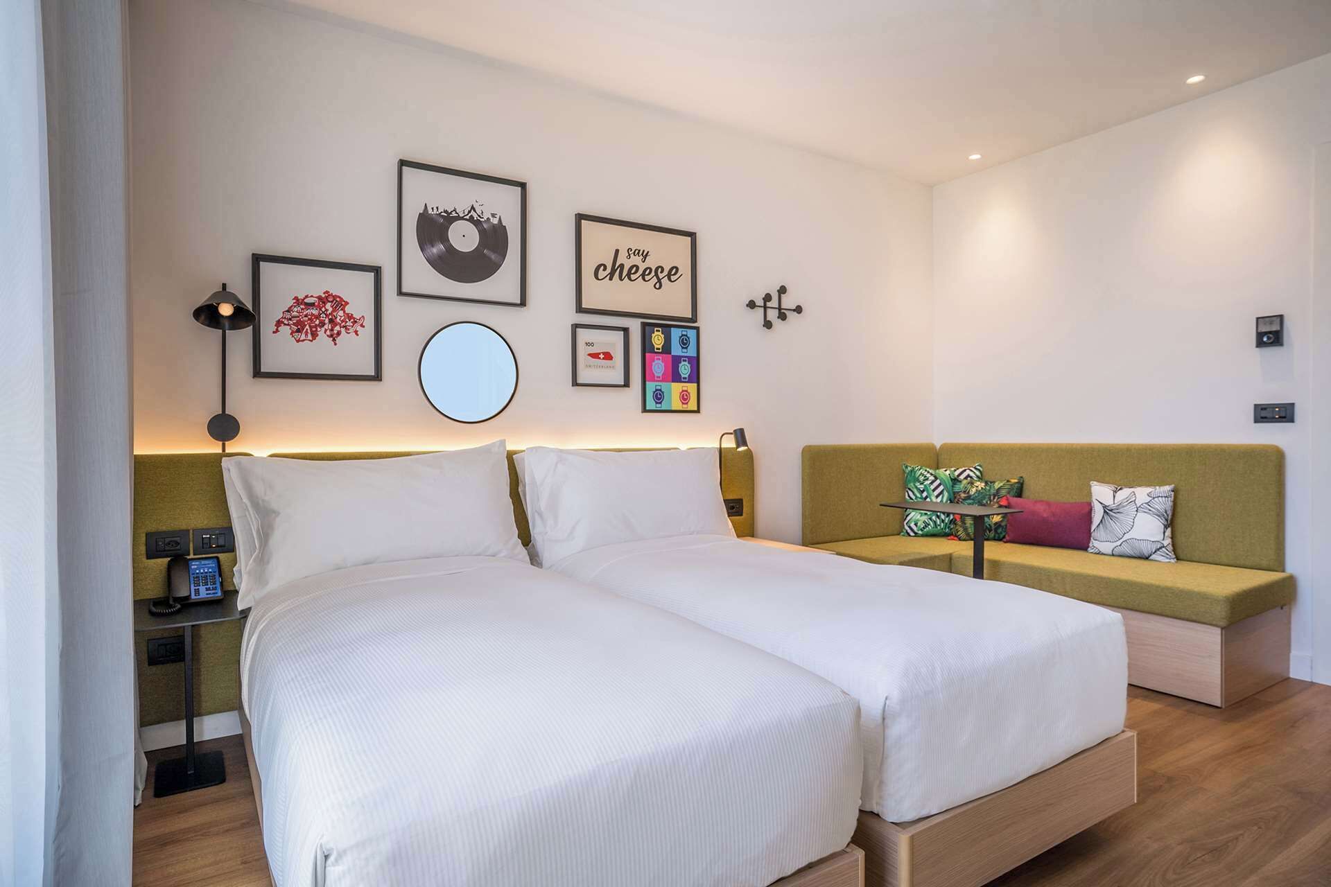 Ein modernes Hotelzimmer mit Doppelbett, einer Couch und einem kleinen Tisch. An den Wänden hängen Bilder in Rahmen, es gibt einen runden Spiegel und eine Lampe auf dem Nachttisch.